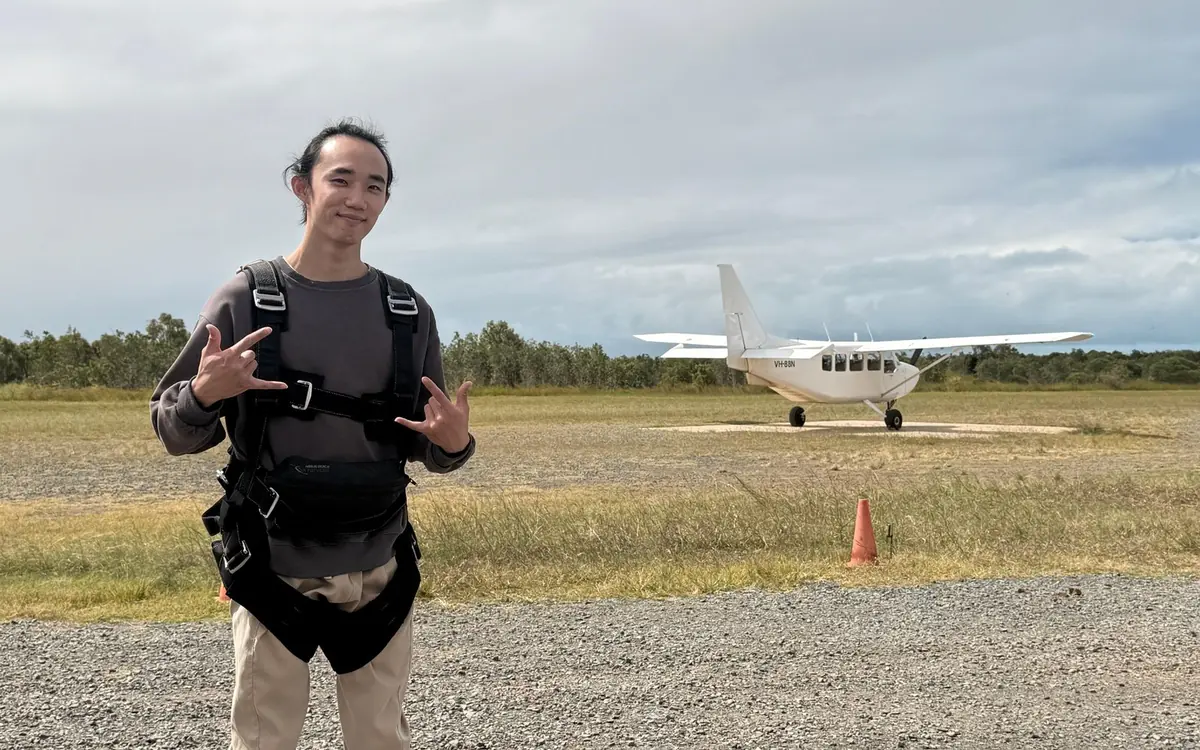 A jumper in a harness on a grass airstrip, a small aircraft parked behind under grey sky